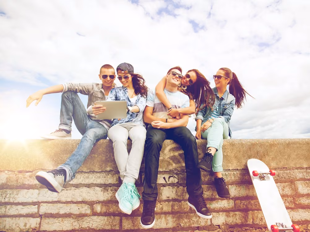 Fünf junge Leute sitzen im Freien und lachen, umgeben von Skateboard und Tablet, bei Tageslicht.