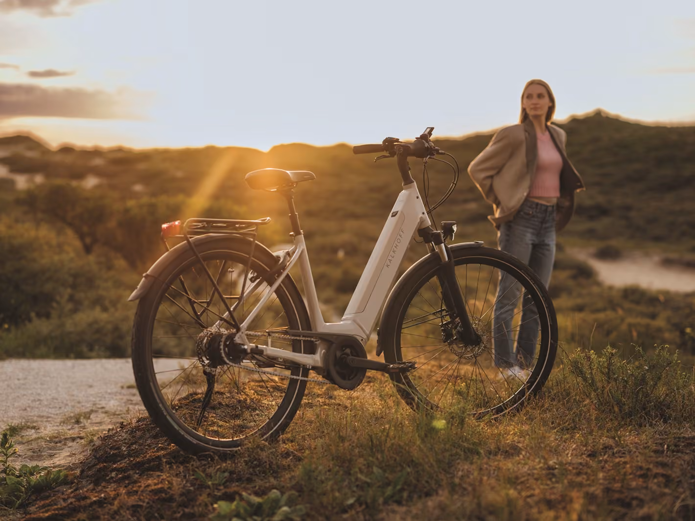 Eine Frau steht bei Sonnenuntergang neben einem weißen E-Bike in einer Landschaft mit Hügeln.