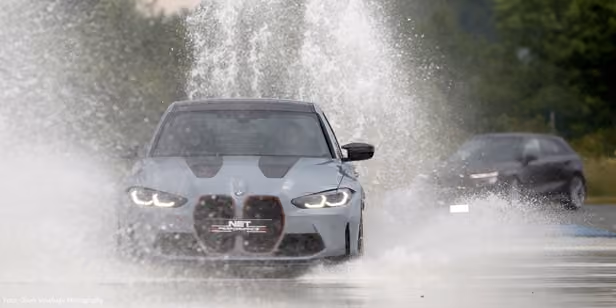 BMW fährt im Sommer 2023 auf nasser Teststrecke, Wasser spritzt hoch, Fokus auf Fahrsicherheit.