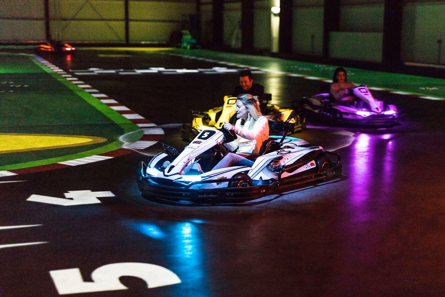 Drei Personen fahren nachts in einer Halle auf bunten Karts auf einer Rennstrecke.