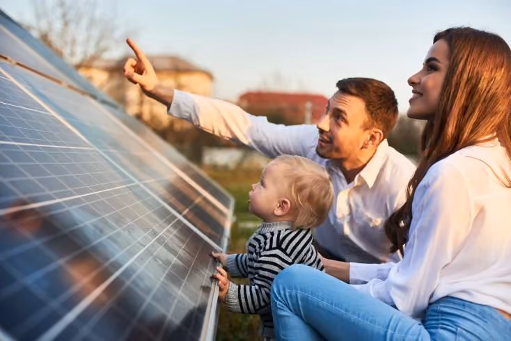 Vater, Mutter und Kind begutachten im Freien ein Solarpanel, Vater zeigt begeistert darauf.