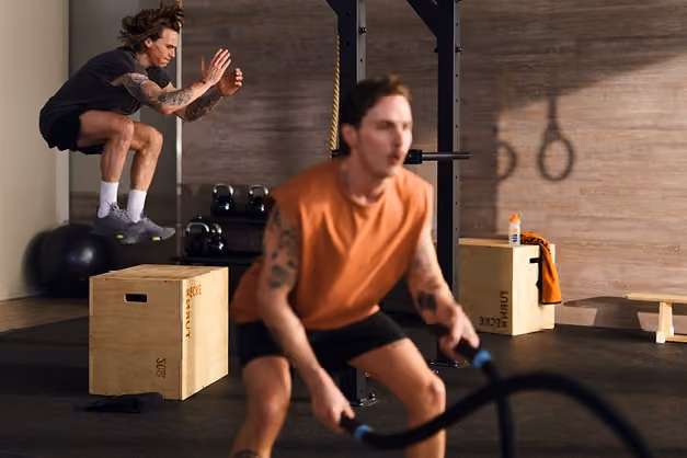 Zwei Männer trainieren im Fitnessstudio, einer macht Sprünge auf eine Holzkiste, der andere nutzt ein Trainingsseil.