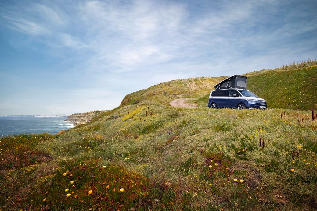 Ein blauer Camper Van steht im Sommer auf einem grünen Hügel an der Küste mit Blick aufs Meer.