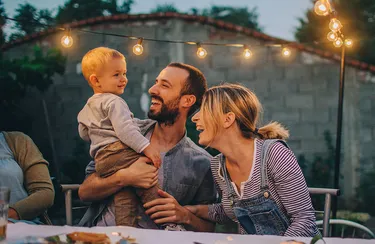 Durch Batteriespeicher kann grüner Strom aus Sonne und Wind immer zur Verfügung stehen. Eine junge Familie feiert abends im Garten bei gemütlicher Beleuchtung.