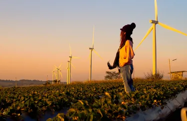 Ein junge Frau steht in einem Feld mit Windenergieanlagen und schaut in die Ferne.