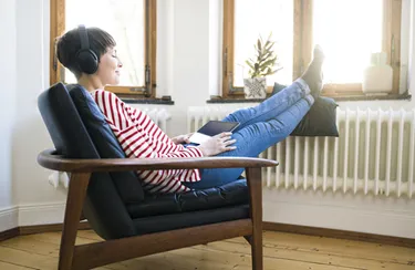 Eine optimal eingestellte Heizung und dichte Fenster helfen beim Energie sparen. Eine junge Frau sitzt in einem Sessel, hört Musik und hat ihre Füße auf der Heizung abgelegt.