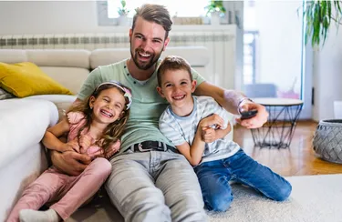 Ein Vater und seine zwei kleinen Kindern sitzen zusammen auf dem Sofa und sehen fern.