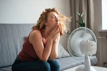 Eine unter sommerlicher Hitze leidende junge Frau sitzt zu Hause vor einem elektrischen Ventilator und kühlt sich mit frischer Luft ab.