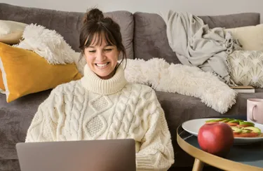 Eine Frau mit einem warmen Rollkragenpullover sitzt auf dem Boden und lehnt sich an ein Sofa. Sie nutzt ihr Notebook und lacht dabei fröhlich.