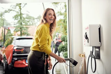 Lachende Frau steht in der Garage und hält den Stecker der wallbe Pro Online Ladebox, um ihr rotes E-Auto zu laden 