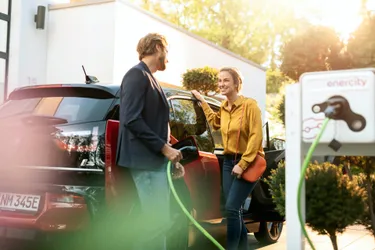 Mann und Frau stehen angelehnt an einem roten E-Auto, rechts steht die wallbe Pro Online Ladesäule. 