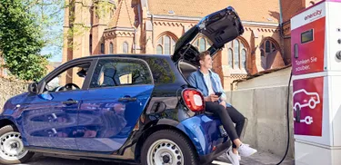 Mann sitzt in Kofferraum seines E-Autos, das an einer Ladebox vor einer Kirche lädt 