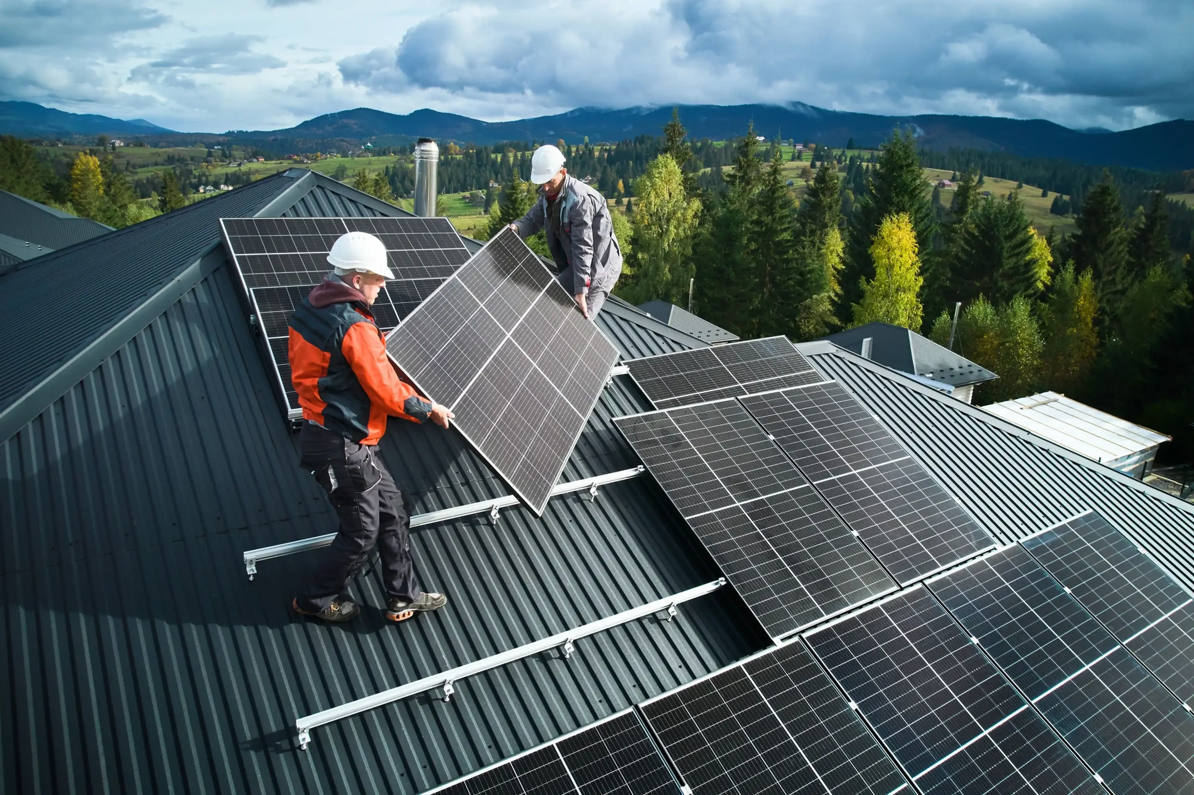 Zwei Monteure in Arbeitskleidung halten ein Solarmodul in den Händen und stehen auf einem Dach, wo schon mehrere Module installiert sind