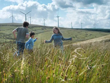 Eine Familie geht durch hohes Gras, während im Hintergrund Windkraftanlagen sichtbar sind.