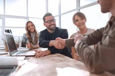 in einem Sitzungsraum schütteln sich zwei Männer die Hände, im Hintergrund sitzen zwei Frau