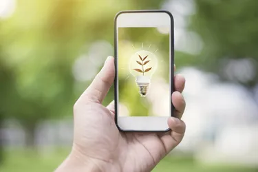 Eine Hand hält einen Handybildschirm hoch auf dem die Illustration einer Glühbirne mit einem leuchtenden grünen Blatt zu sehen ist