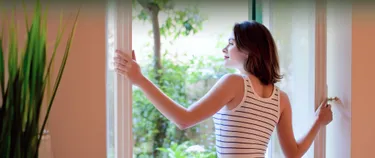 Eine junge Frau steht an einem geöffneten Fenster im Altbau und genießt die Frischluft