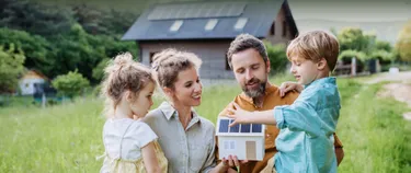 Eine Familie steht im Garten und schaut sich das Modell eines Hauses mit einer PV-Anlage auf dem Dach an.
