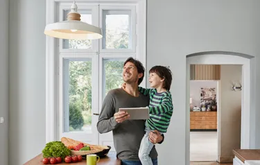 Ein Mann mit einem Tablet in der Hand und einem kleinen Jungen auf dem Arm steht in einer Küche und guckt Richtung Deckenlampe