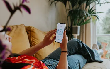 Eine Frau liegt auf einem Sofa und reguliert per App auf einem Handy die in ihrer Wohnung herrschende Raumtemperatur