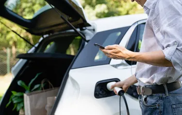 Ein Mann lädt sein Elektroauto und hält dabei ein Smartphone in der Hand