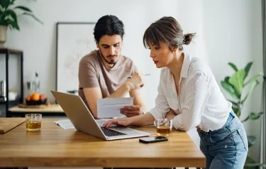 Ein Mann lehnt an einem Hochtisch und studiert eine Rechnung, während eine Frau an einem Laptop arbeitet