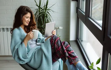 Eine Frau sitzt in eine Wolldecke gekuschelt auf einem Stuhl neben einem Heizkörper und schaut in ein Tablet 