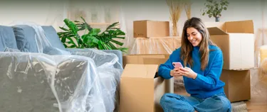 Eine junge Frau sitzt mit dem Smartphone auf dem Boden zwischen Umzugskartons und wechselt ihren Stromanbieter