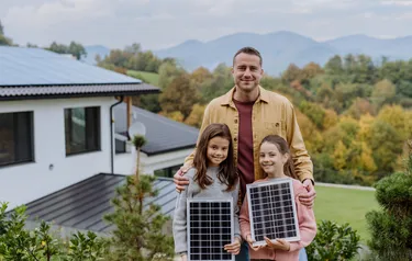 Ein Vater steht mit seinen zwei Töchtern vor ihrem mit PV-Anlagen bestückten Haus