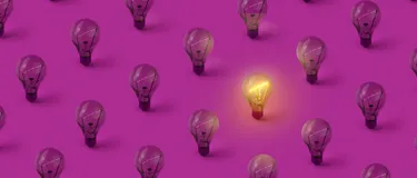 Dieses Bild zeigt viele Glühbirnen auf einem lilafarbigen Untergrund. Eine von den Glühbirnen leuchtet, alle anderen sind aus.
