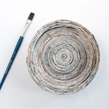 Das Bild zeigt eine Schale aus gefaltetem und aufgerolltem Zeitungspapier sowie einen Pinsel.