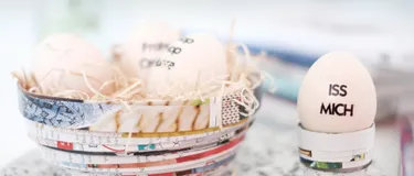 Das Bild zeigt ein mit drei Eiern gefülltes Osternest aus gefaltetem und zusammengerolltem Zeitschriftenpapier sowie einen Eierbecher aus dem gleichen Material, in dem ein Ei steckt.