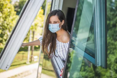 Hygienekonzept. Dieses Bild zeigt eine junge Frau, die eine medizinische Maske auf hat und gerade ein Fenser öffnet.