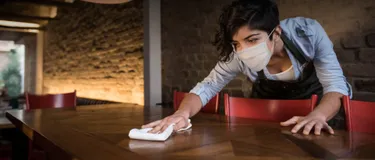 Dieses Bild zeigt eine junge Frau mit schwarzen kurzen Haaren, die eine Maske trägt und einen Tisch putzt.