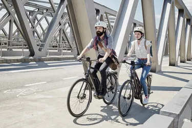 Das Bild zeigt ein junges Paar, das mit Pedelecs über eine Brücke in einer urbanen Umgebung fährt.
