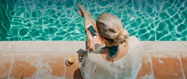 Handy, Laptop oder Tablet überhitzt? Das hilft! Das Bild zeigt eine Frau, die im Hochsommer am Pool sitzt und ihr Smartphone bedient.