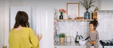 Stromverbrauch beim Kühlschrank senken: Das Bild zeigt eine Mutter, die die Tür eines Kühlschrankes öffnet, während ihr Kind dabei auf der Arbeitsplatte sitzt.
