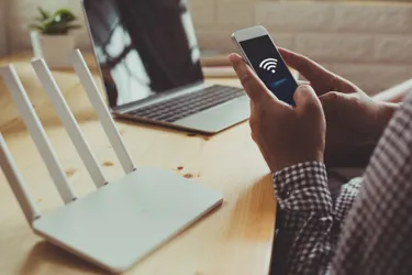 Eine Person hält ihr Handy, davor steht ein Laptop und ein Router.