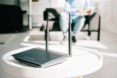 Ein WLAN-Router steht auf einem kleinen Tisch, im Hintergrund sitzt eine Person auf dem Sofa.