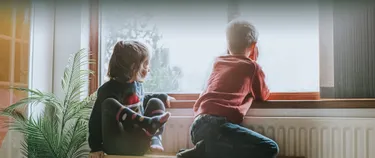 Zwei Kinder sitzen an der Heizung vor einem Fenster und sehen hinaus.