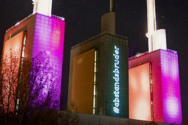 Dieses Bild zeigt das Kraftwerk von Linden bei Nacht. Zwei der drei Türme leuchten, während der mittlere dunkel ist. Auf seiner Vorderseite ist in Leuchtschrift "Abstandsbruder" zu erkennen.