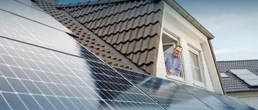 Vorreiter: Energie-Selbstversorger. Dieses Bild zeigt einen Mann, der aus einem Dachfenster schaut. Auf dem Dach ist eine Solaranlage installiert.