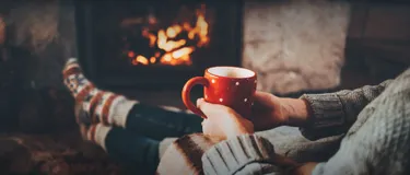 Heizen mit Holz: Wie gut ist es wirklich für das Klima? Tipps für nachhaltiges Heizen mit Holz. Das Bild zeigt eine junge Frau, die gemütlich mit einer Tasse Kaffee vor dem Kamin sitzt.