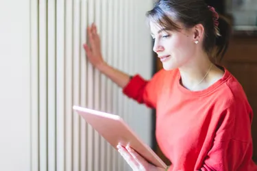 Junge Frau mit einem Tablet in der linken Hand. Mit der rechten Hand berührt die Frau einen Heizkörper.