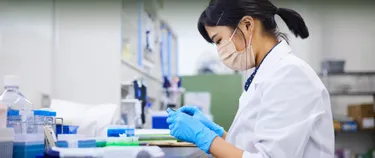 Eine Frau mit Mundschutz und Handschuhen arbeitet mit Gerätschaften in einer Laborumgebung.