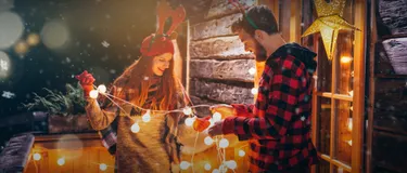 Nachhaltige Weihnachtsbeleuchtung. Das Bild zeigt eine Frau und einen Mann, die gemeinsam eine Lichterkette mit Kugeln festhalten. Die Menschen stehen vor einem Holzhaus und sind weihnachtlich gekleidet.