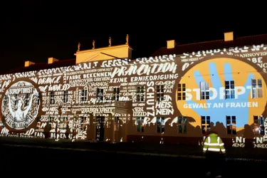 Dieses Bild zeigt das Schloss Herrenhausen, welches orange angestrahlt wurde. Neben der Botschaft "Stoppt Gewalt gegen Frauen" werden auch andere Begriffe wie "Prävention", "Schutz" oder "Farbe bekennen" an die Fassade projiziert.