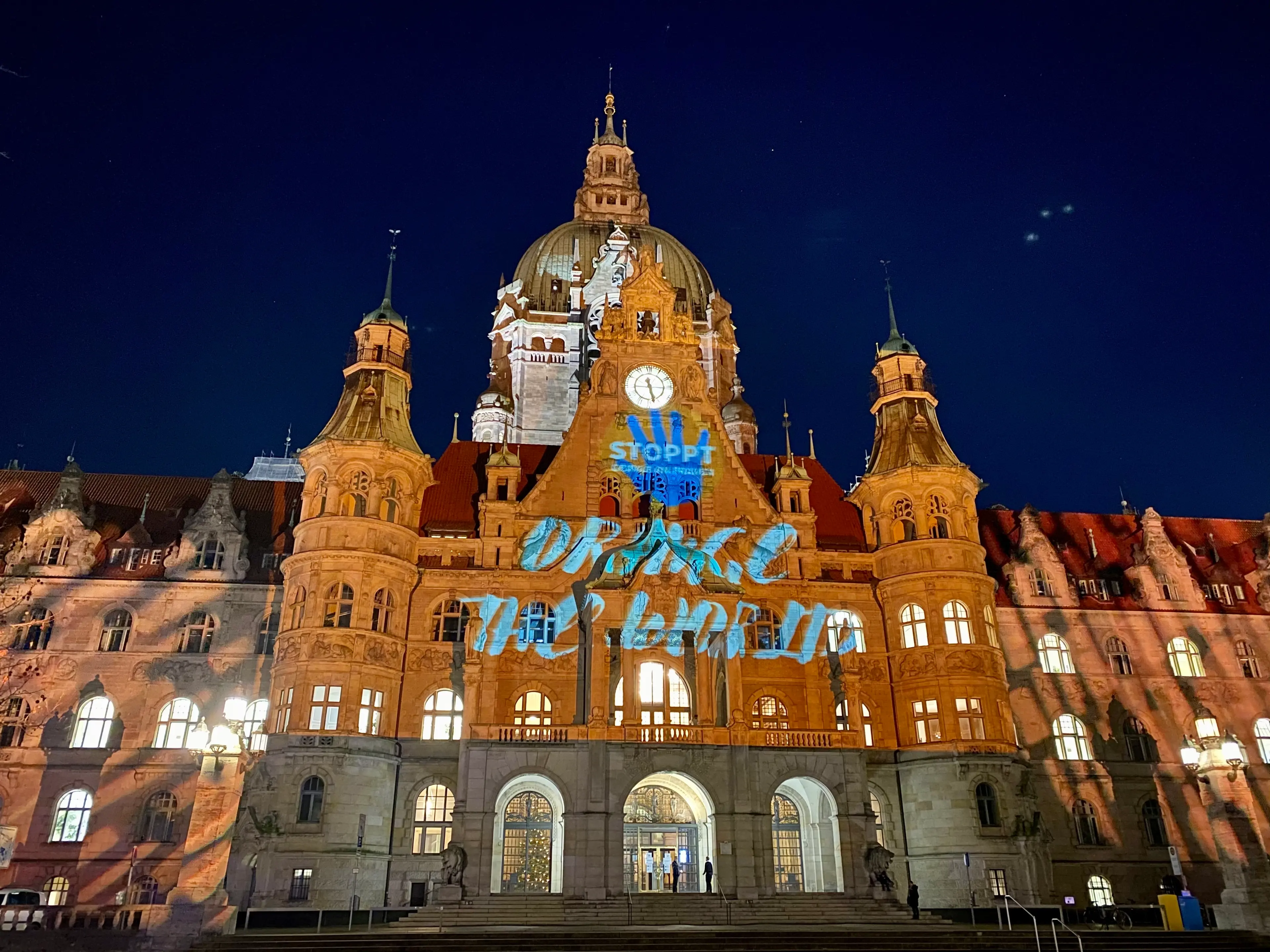 Dieses Bild zeigt das neue Rathaus Hannovers, welches orange angestrahlt wurde. Zudem wurde der Schriftzug "Stoppt Gewalt gegen Frauen" in blau auf das Gebäude projiziert. 