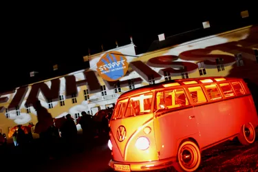 Dieses Bild zeigt einen alten Bulli, der orange angestrahlt wird. Im Hintergrund sieht man verschiedene Menschen und die Botschaft "Stoppt Gewalt gegen Frauen".