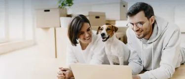 Energieeffizientes Wohnen. Dieses Bild zeigt einen Mann und eine Frau am Laptop. Zwischen ihnen sitzt ein Hund.